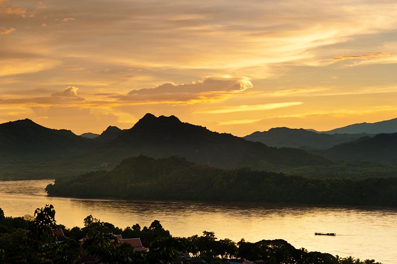 Fotografía de un atardecer en el río Mekong. En una margen del río se ven casa y vegetación y en la otra montañas. Una pequeña barca. El cielo tiene multitud de tonos amarillos y naranjas.