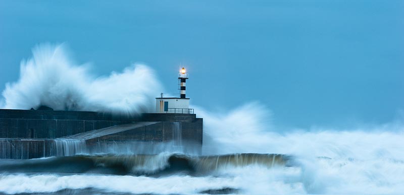 Fotografía del faro de San Estaban de Pravia en pleno temporal con las olas chocando contra el muro de contención del puerto y saltando por encima