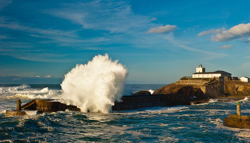 Fotografía del faro de Tapia de Casariego con una ola rompiendo contra el muro de protección del puerto. El mar está muy revuelto. Al fondo se ven olas y nubes de tormenta