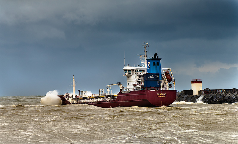 Fotografía de un barco carguero entrando por la ría del río Adour durante una tormenta y temporal en el mar. El mar está muy agitado, en tonos marrones y blanco, las olas golpean el barco y el muro de contención donde la baliza roja o de estribor se mantiene firme.