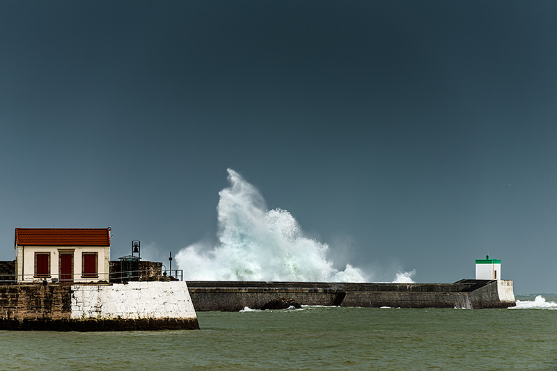 Fotografía del puerto de Saint Jean de Luz, con el mar de color verde esmeralda, un cielo gris plomizo por la tormenta y una ola que rompe contra el muro de contención del puerto y que salta por encima. Al fondo del muro de contención del puerto se ve la baliza verde y en primer término un edificio de color rojo que contrasta con todos los colores del resto de la fotogra´fia