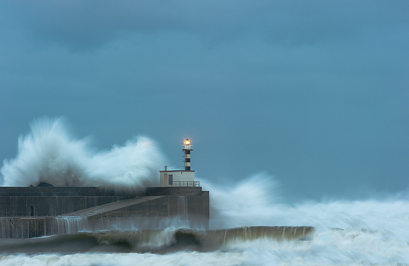 Fotografía del faro de San Estaban de Pravia en pleno temporal con las olas chocando contra el muro de contención del puerto y saltando por encima
