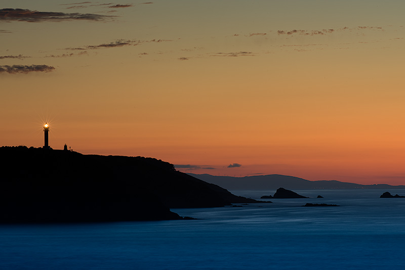 Fotografía del faro de San Agustín en Ortiguera, Asturias. La linterna está encendida y se encuentra en la parte izquierda de la fotografia. El acantilado hacia el mar en tonos grises y negros, el mar en tonos azules cobaltos y el cielo todo tipo de tonos ocres. Varias rocas sobresalen del agua enfrente de la costa