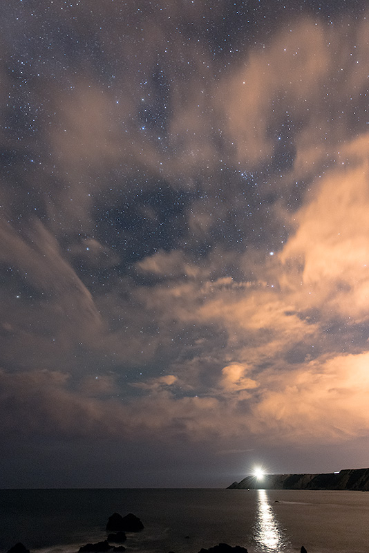 Fotografía nocturna del faro encendido del cabo Vidio y de la costa de Cudillero con la playa del Silencia en primer plano.