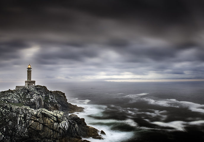 Fotografía del faro de Punta Nariga. en la parte de la izquierda se ve el faro encendido y parte de los acantilados de costa. En la parte de la derecha el mar en tonos azules y grises.