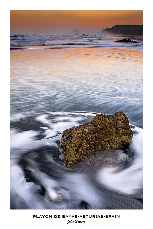 Fotografía del Playón de Bayas en Castrillón Asturias, España. Una roca en la playa a la que le rodea una ola que retrocede. Al fondo el perfil de la costa con los colores ocres de un atardecer.