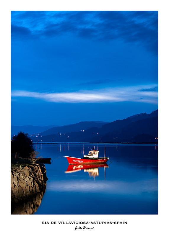 Fotografia de la ría de Villaviciosa en Asturias, España, al atardecer. En primer plano se ve una barca de faenar de color rojo. El cielo y el agua tienen multitud de colores azules.