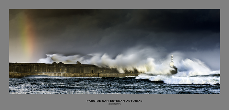Faro de San Esteban de Pravia en pleno temporal con una ola rompiendo contra el muro de contención del puerto y la linterna del faro y como la ola entra por la parte más abierta. El muro se ve claramente y como el agua arrolla. El color de mar es de un azul metalizado y el del cielo tiene muchos tonos grises y azules. En la parte de la izquierda se difumina un arco iris.
